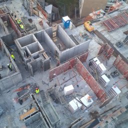 PRZEDSIĘBIORSTWO BUDOWLANE IKC SPÓŁKA Z OGRANICZONĄ ODPOWIEDZIALNOŚCIĄ - Budowa fundamentów budynku z betonu, widok z góry. Widoczne szalunki, zbrojenia, pracownicy w żółtych kamizelkach i mała koparka. Na drugim planie budynki i zieleń.