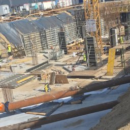 PRZEDSIĘBIORSTWO BUDOWLANE IKC SPÓŁKA Z OGRANICZONĄ ODPOWIEDZIALNOŚCIĄ - Budowa fundamentów z widocznymi szalunkami, zbrojeniami i rurami. W tle dźwig budowlany i budynek. Prace ziemne w toku, widoczne wykopy i elementy konstrukcyjne.