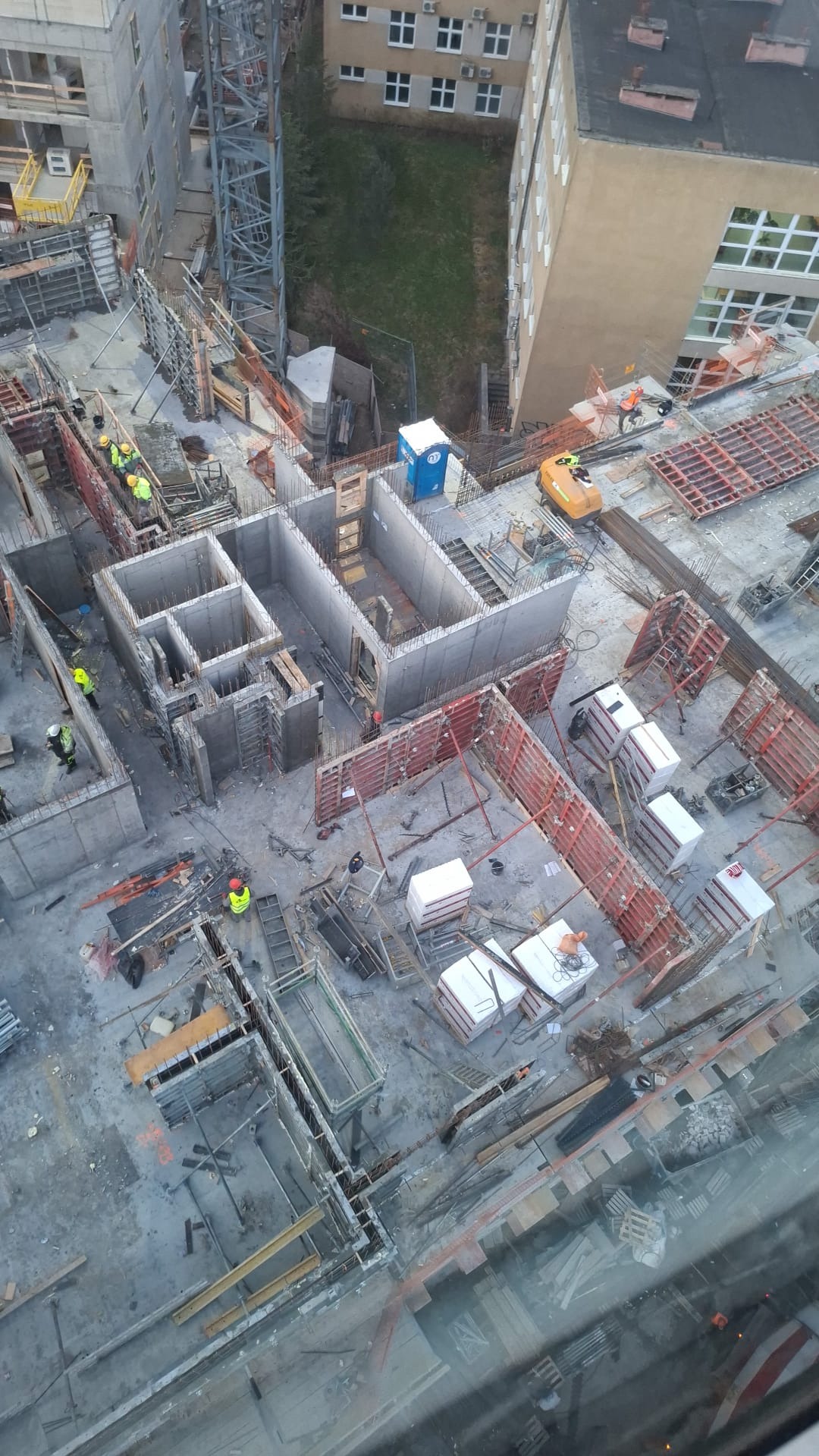 Budowa fundamentów budynku z betonu, widok z góry. Widoczne szalunki, zbrojenia, pracownicy w żółtych kamizelkach i mała koparka. Na drugim planie budynki i zieleń.