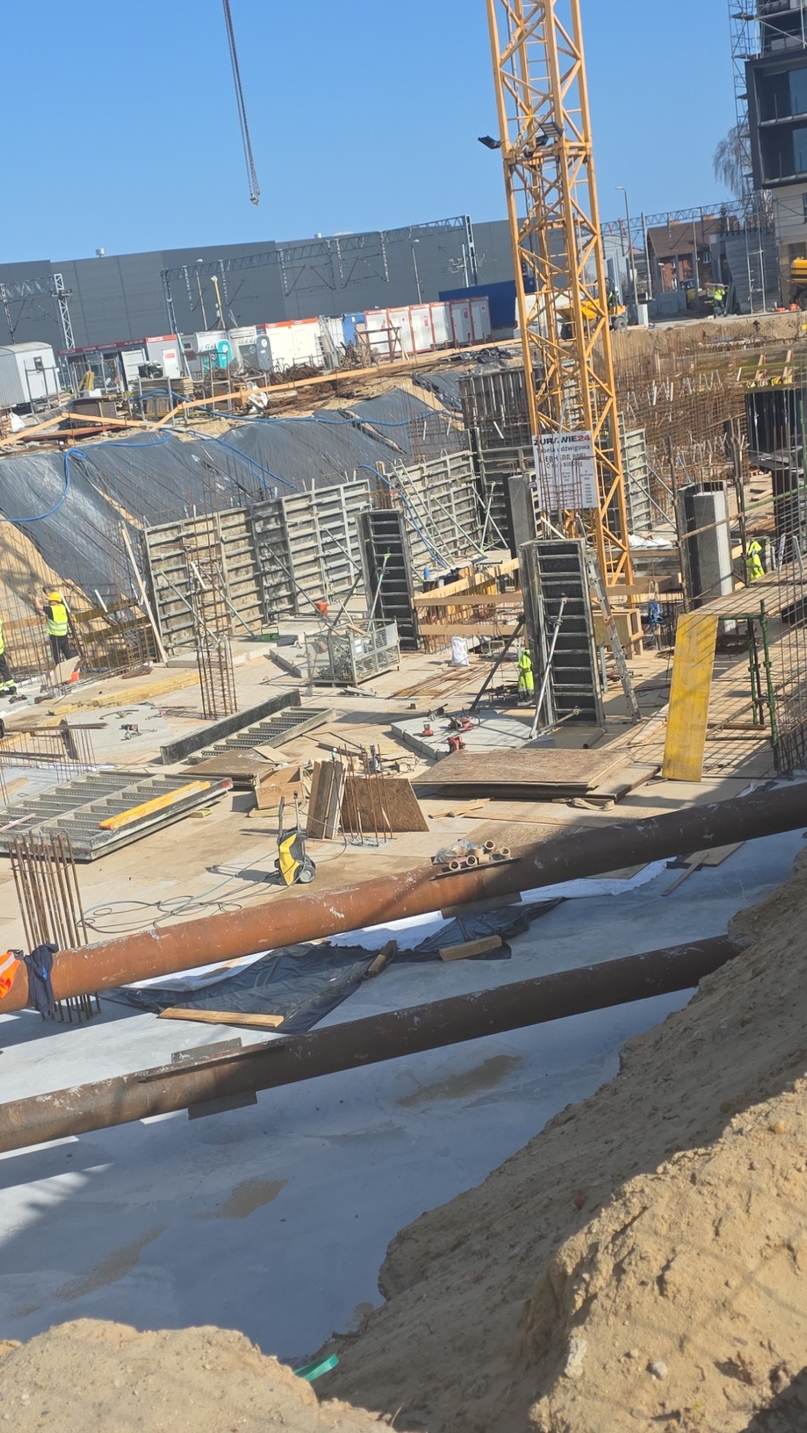 Budowa fundamentów z widocznymi szalunkami, zbrojeniami i rurami. W tle dźwig budowlany i budynek. Prace ziemne w toku, widoczne wykopy i elementy konstrukcyjne.