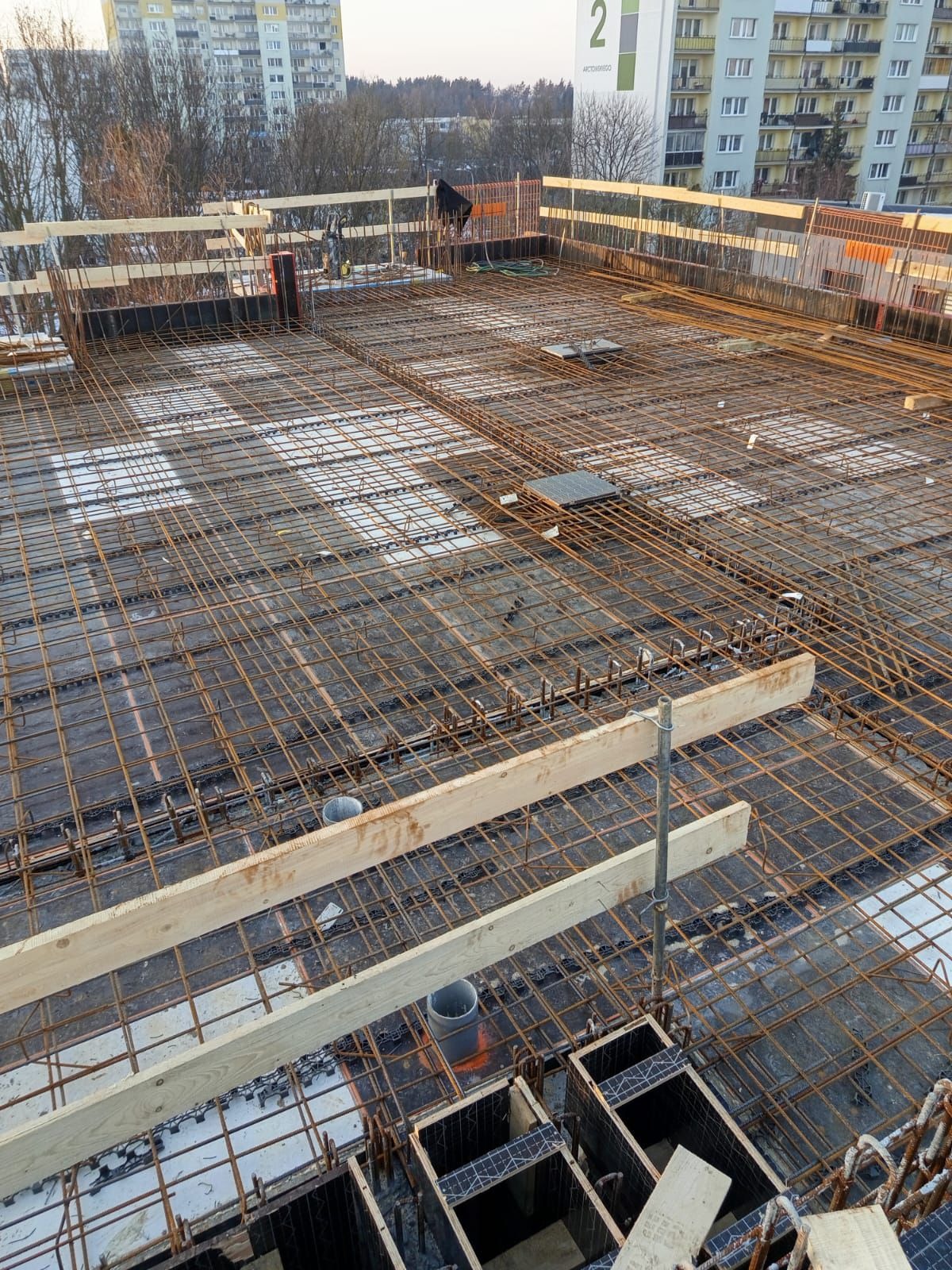 Siatka zbrojeniowa na stropie budynku w trakcie budowy, widok z góry. Elementy szalunkowe i drewniane podpory. W tle budynki mieszkalne.