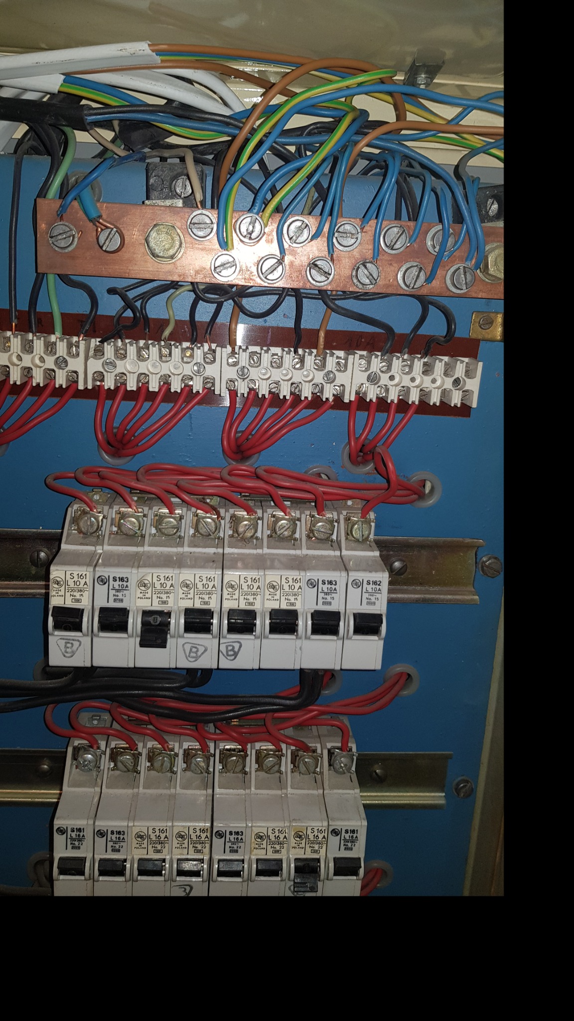 Rozdzielnica elektryczna z widocznymi bezpiecznikami, listwami zaciskowymi i przewodami w różnych kolorach, zamontowana w niebieskiej obudowie.