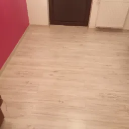 Jasne panele podłogowe imitujące drewno, ułożone w pokoju z różową ścianą i brązowymi drzwiami, widoczny biały grzejnik.