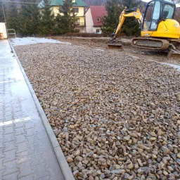 Żwirowe podłoże na placu budowy, z żółtą koparką JCB w tle, gotowe pod dalsze prace.