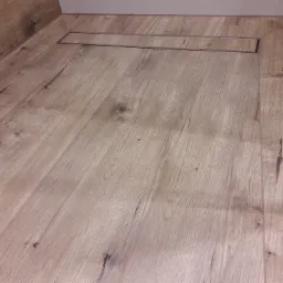 Podłoga w łazience wyłożona panelami imitującymi drewno, z liniowym odpływem prysznicowym w kolorze stali nierdzewnej i fragmentem szklanej kabiny prysznicowej.