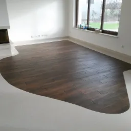 Wykończona podłoga w salonie z ciemnymi panelami imitującymi drewno ułożonymi w nieregularny kształt na jasnych, błyszczących płytkach, widoczny kominek i okno z parapetem.