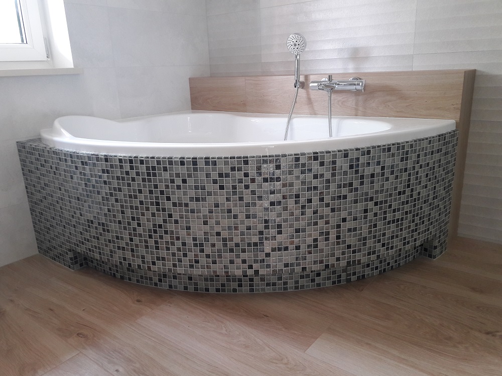 Biała narożna wanna obudowana mozaiką w odcieniach szarości, z drewnianym panelem za baterią i słuchawką prysznicową, na podłodze z paneli imitujących drewno.