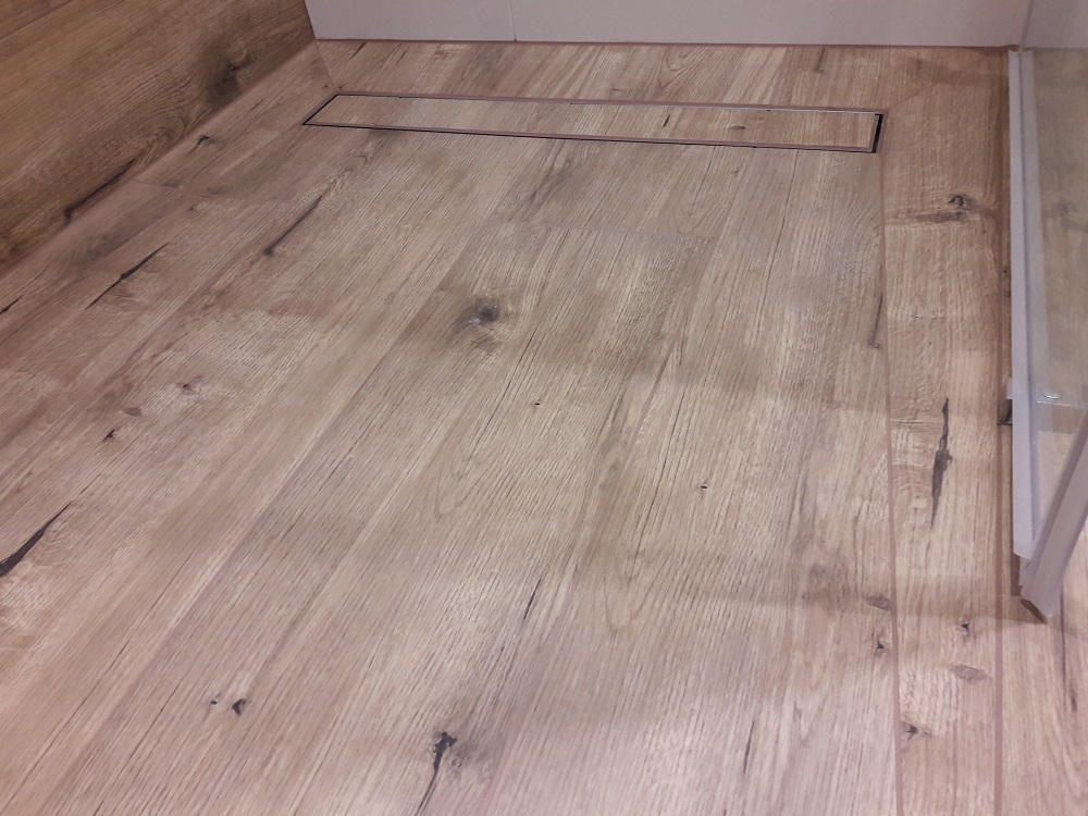 Podłoga w łazience wyłożona panelami imitującymi drewno, z liniowym odpływem prysznicowym w kolorze stali nierdzewnej i fragmentem szklanej kabiny prysznicowej.