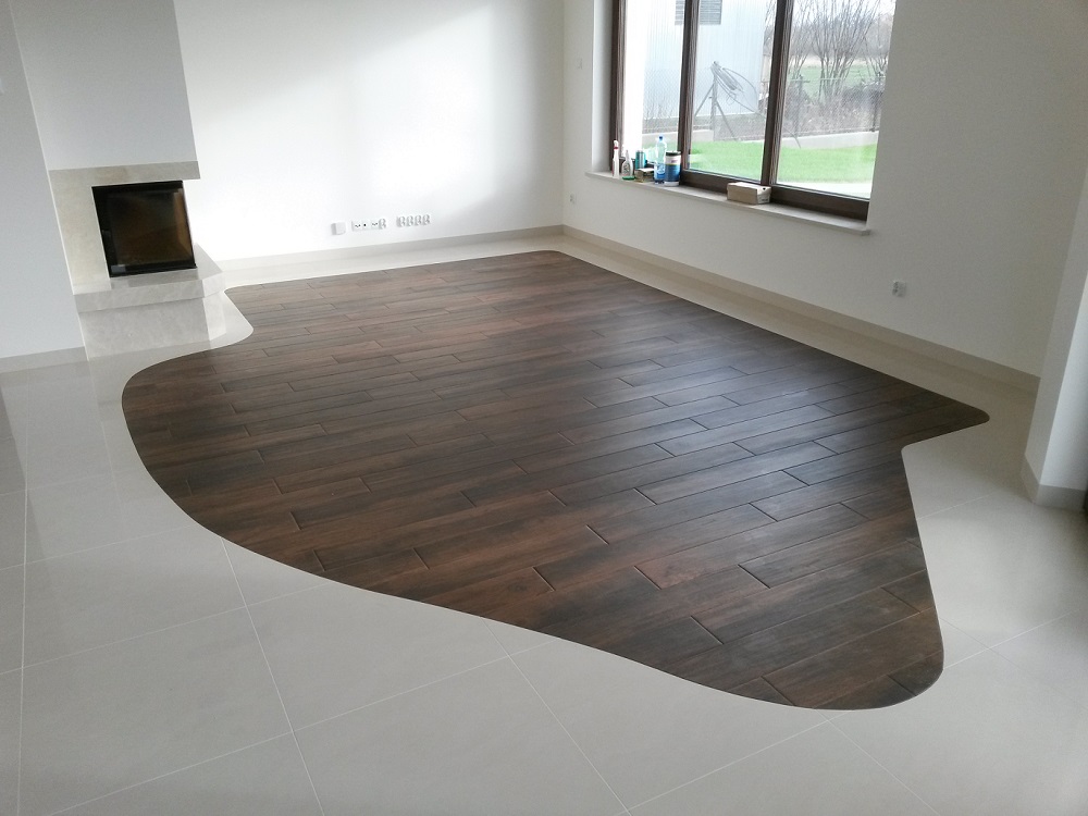 Wykończona podłoga w salonie z ciemnymi panelami imitującymi drewno ułożonymi w nieregularny kształt na jasnych, błyszczących płytkach, widoczny kominek i okno z parapetem.