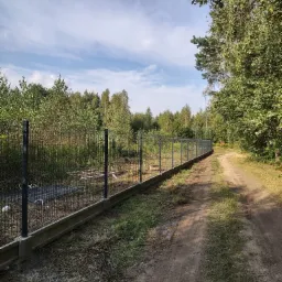 Długi fragment panelowego ogrodzenia na betonowej podmurówce, biegnący wzdłuż polnej drogi, z lasem w tle i błękitnym niebem z chmurami nad nim.