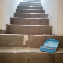 Schody wyłożone szarymi płytkami z metalowymi listwami, widoczna niebieska skrzynka z narzędziami, białe ściany i jasne światło u góry schodów.