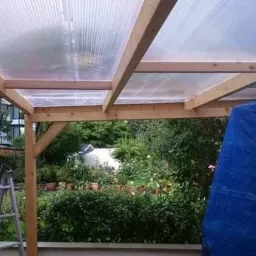 Zadaszenie tarasu z przezroczystego poliwęglanu falistego na drewnianej konstrukcji, widok na ogród, drabina oparta o ścianę, niebieska plandeka w rogu.