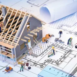 Model domu w trakcie budowy z miniaturowymi figurkami robotników na planie architektonicznym, ukazujący etap wznoszenia konstrukcji z elementami murowanymi i drewnianą więźbą dachową.