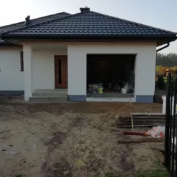 Nowoczesny dom jednorodzinny z białymi ścianami elewacyjnymi, czarną dachówką i otwartym garażem, w którym widoczne są puszki z farbą i narzędzia malarskie, teren wokół domu w trakcie prac...