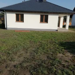 Nowoczesny, parterowy dom z białą elewacją, brązowymi oknami i ciemną, metalową dachówką w słoneczny dzień.