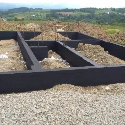 Zabezpieczone, pomalowane na czarno fundamenty domu w trakcie budowy, widoczne wystające pręty zbrojeniowe, teren wokół usypany ziemią i gruzem, w tle krajobraz wiejski.