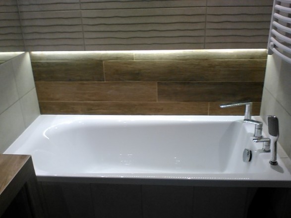 Biała wanna z chromowaną armaturą i deszczownicą, podświetlona listwą LED, otoczona płytkami imitującymi drewno i szarymi płytkami z falistym wzorem.