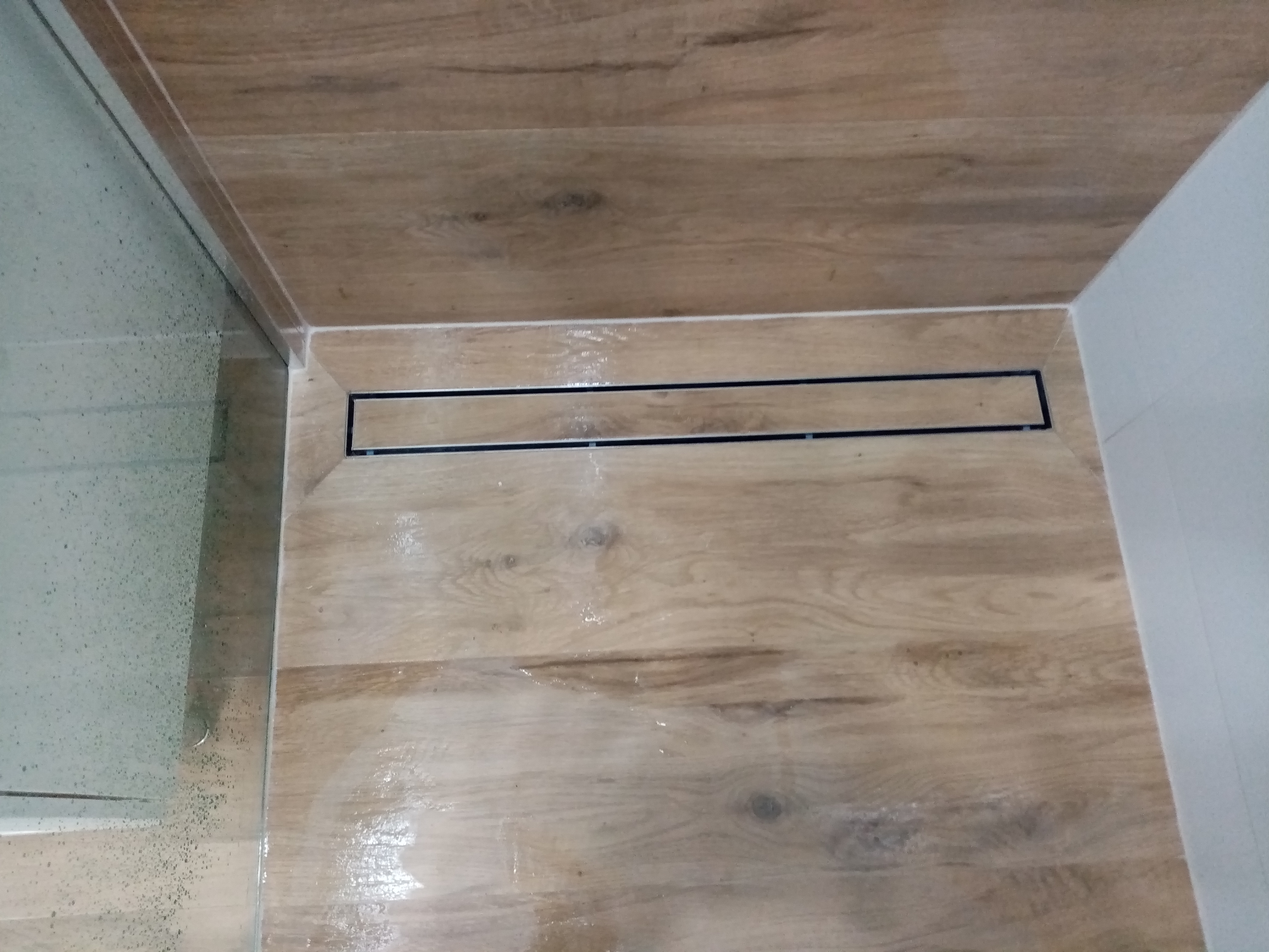 Wykończona kabina prysznicowa z odpływem liniowym i płytkami imitującymi drewno, fuga w kolorze białym.