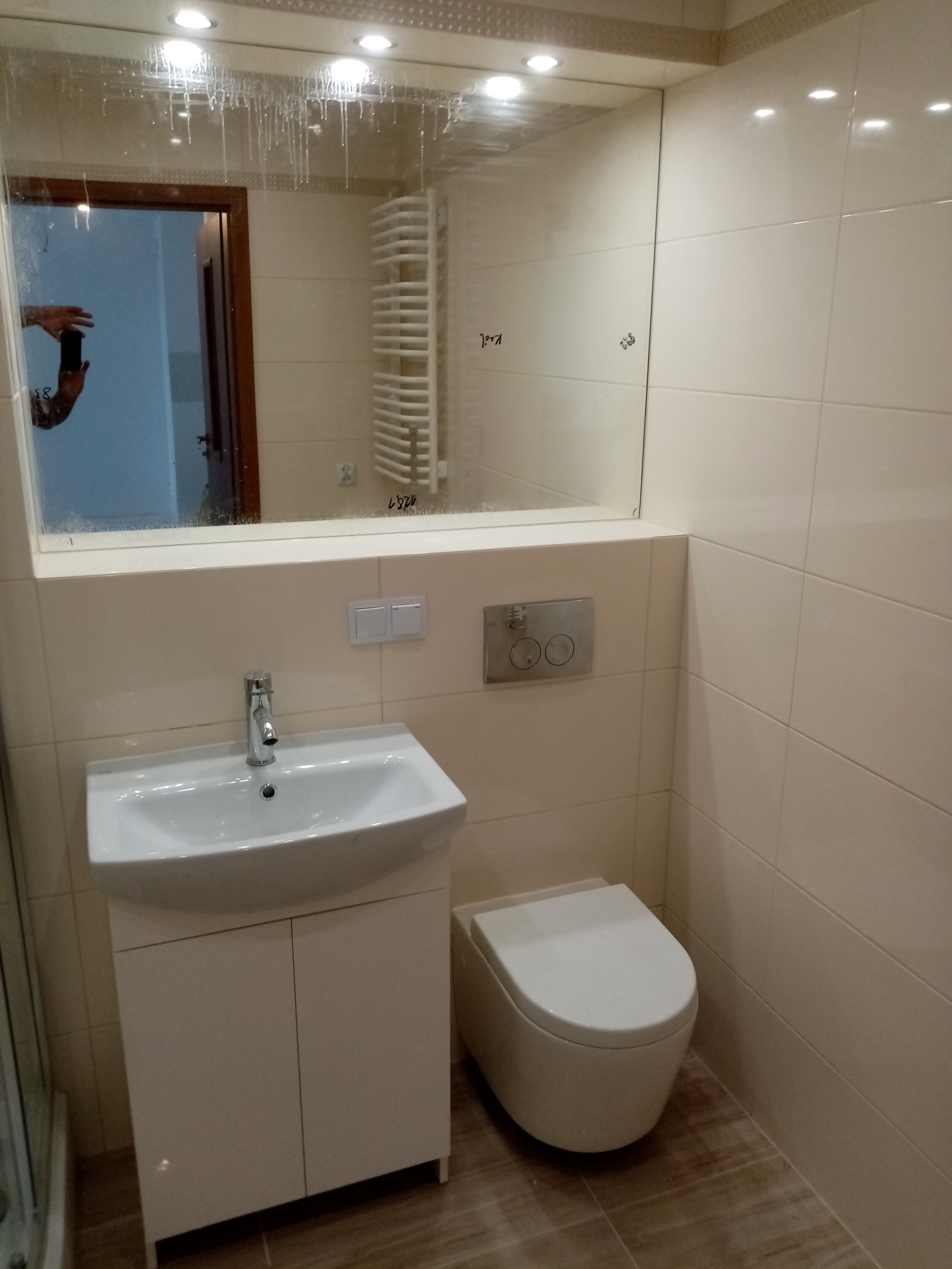 Jasna łazienka z umywalką na szafce, podwieszaną toaletą i dużym lustrem z widocznymi śladami po czyszczeniu na powierzchni.