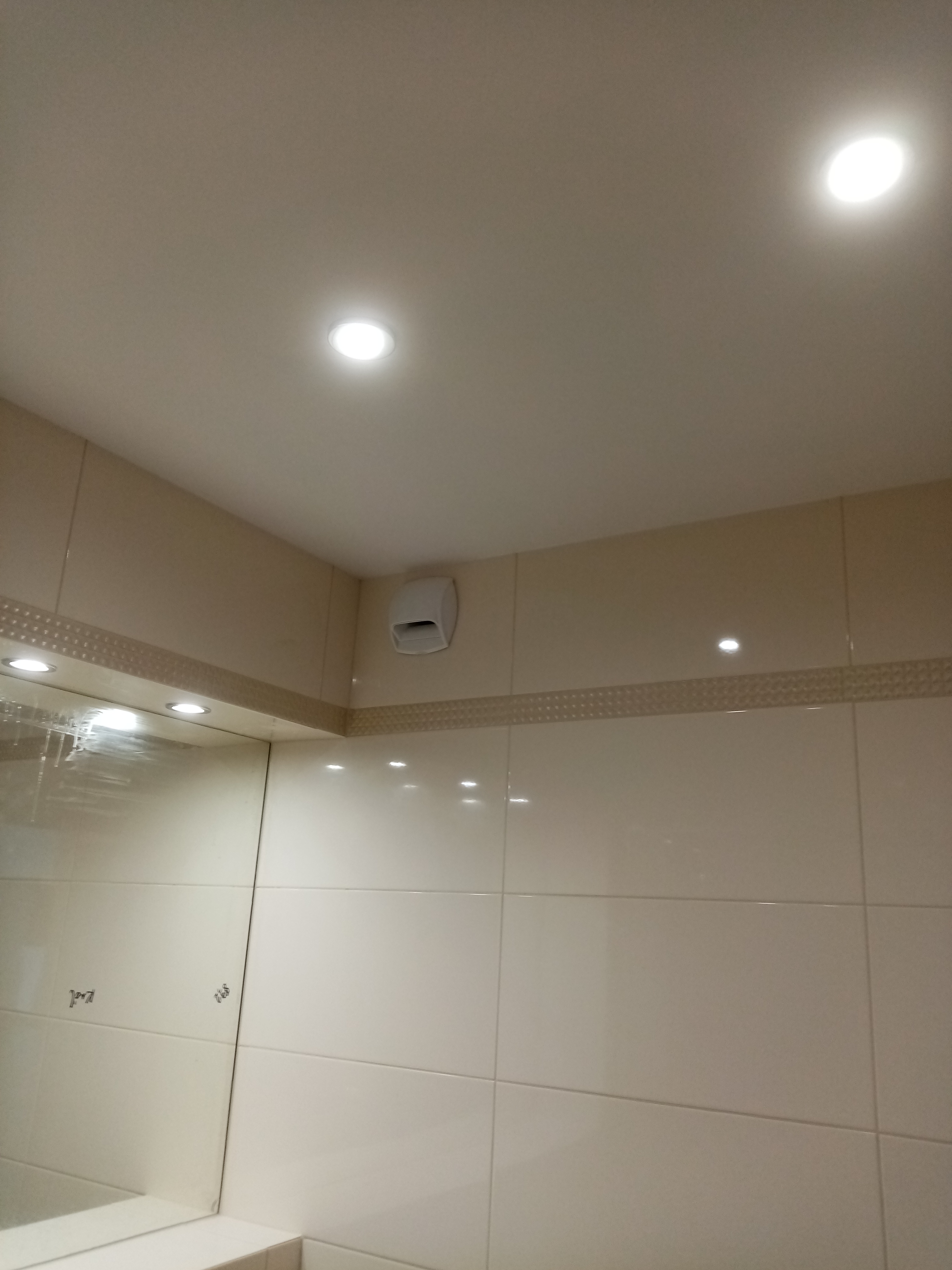 Jasne, beżowe płytki ceramiczne na ścianach łazienki, z dekoracyjnym pasem płytek i widocznym wentylatorem oraz oświetleniem punktowym.