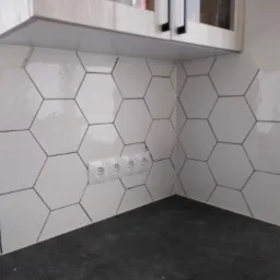 Białe, heksagonalne płytki ceramiczne z czarnymi fugami na ścianie w kuchni, nad blatem. Widoczne gniazdka elektryczne.