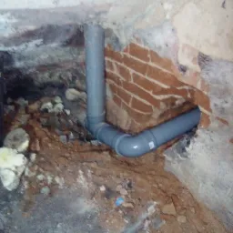 Szara rura kanalizacyjna z kolankami, zamontowana w surowym otoczeniu z cegły i gruzu, widoczne ślady pianki montażowej.