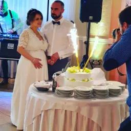 Uśmiechnięta para młoda w otoczeniu tortu weselnego z fontannami iskier, DJ w tle i fotograf robiący zdjęcie.