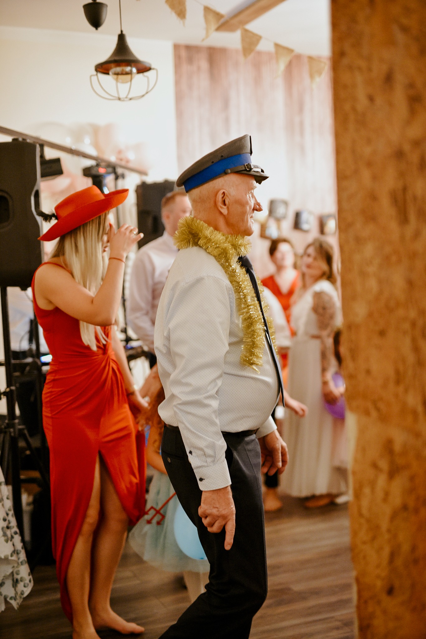 Uczestnicy imprezy: mężczyzna w czapce i złotym łańcuchu, kobieta w czerwonej sukience obok nagłośnienia, w tle inni goście.