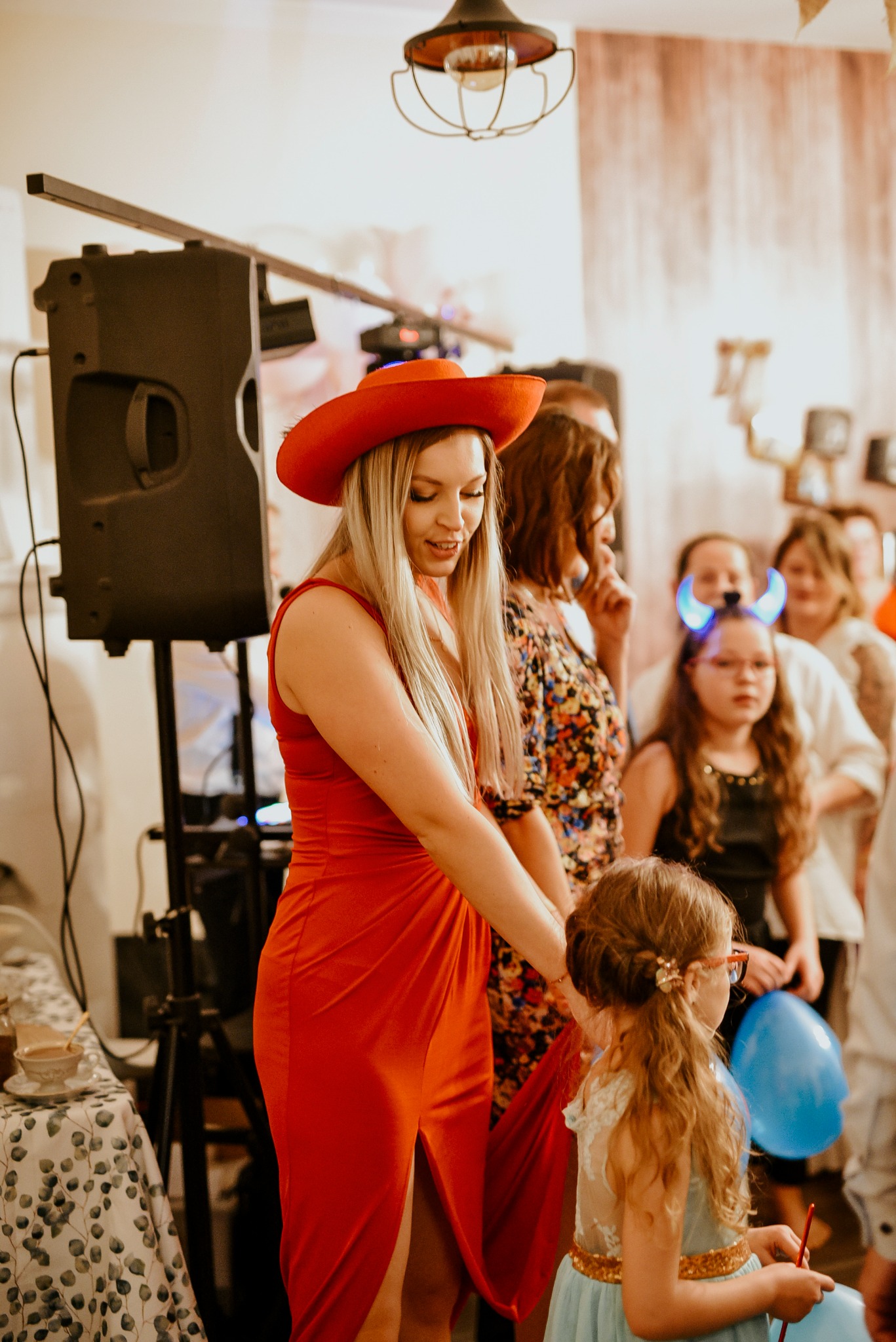 Kobieta w czerwonej sukience i kapeluszu, prawdopodobnie animatorka, w trakcie interakcji z małą dziewczynką na przyjęciu, w tle widoczny głośnik i grupa osób.