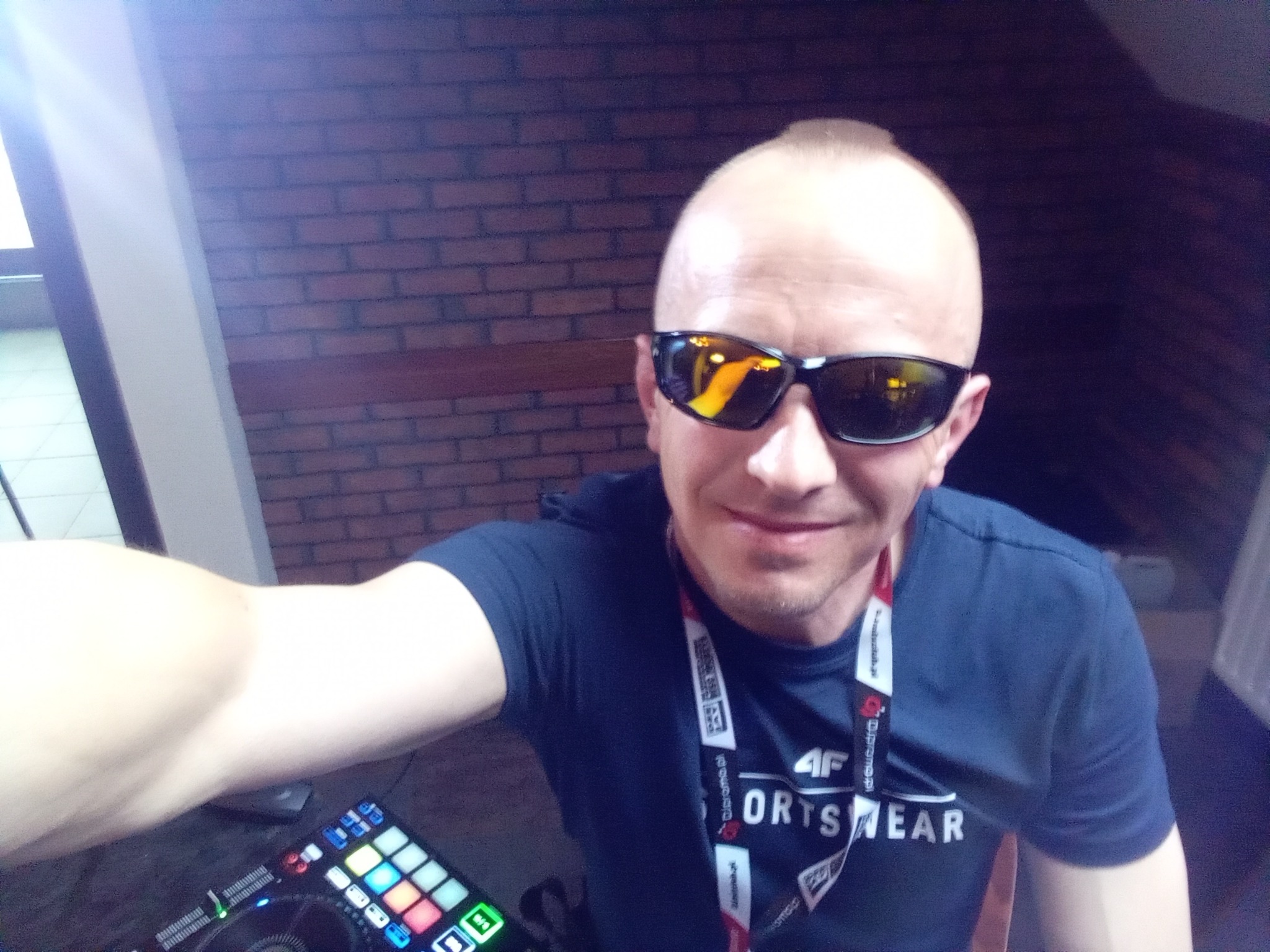 DJ robi selfie z mikserem na tle ceglanej ściany, nosi ciemne okulary i koszulkę z logo 4F.