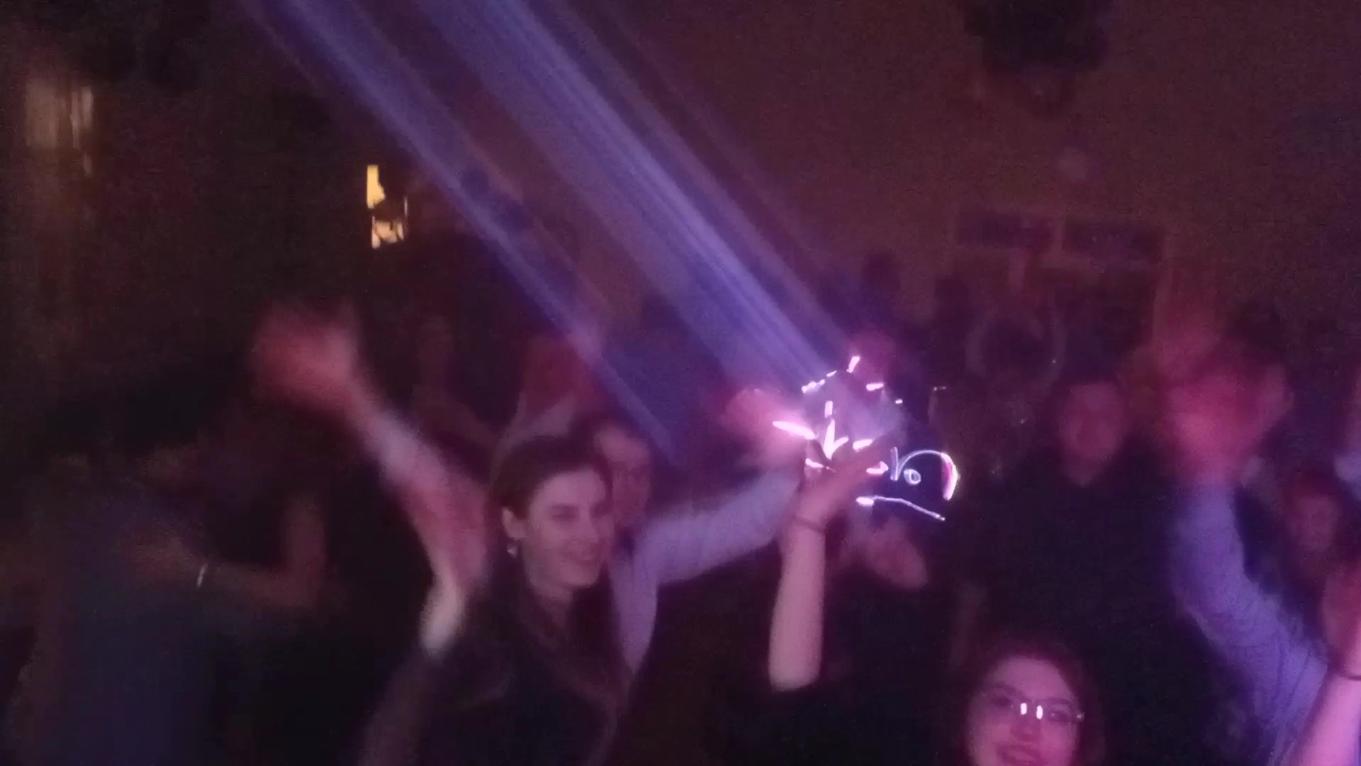 Rozmazane ujęcie z imprezy z tańczącymi ludźmi, oświetlonej fioletowymi reflektorami, z osobą w czapce z ledowym napisem