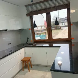 Nowoczesna kuchnia z białymi szafkami, czarnym blatem i drewnianymi żaluzjami w oknie, widoczne czarne lampy wiszące i drewniany stołek.