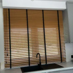 Drewniane żaluzje w kolorze miodowym z czarnymi taśmami w nowoczesnej kuchni z szarymi szafkami i czarnym zlewem. Widoczne niedokończone oświetlenie sufitowe.