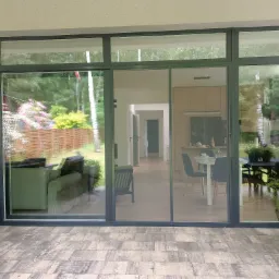 Przesuwne okno tarasowe z szarymi ramami, widok na ogród i wnętrze domu, na pierwszym planie patio z brukowaną podłogą.