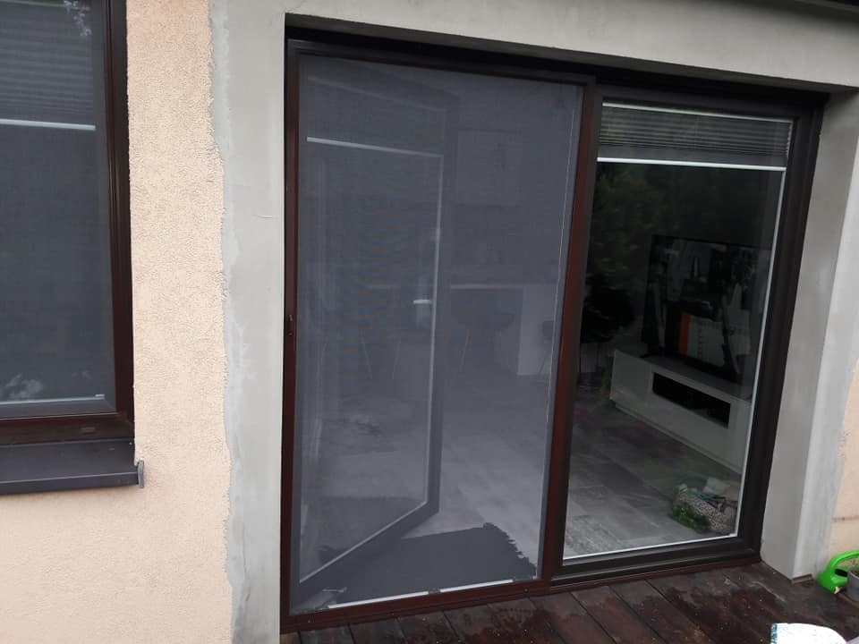 Brązowa moskitiera ramkowa zamontowana w drzwiach tarasowych, widok na wnętrze domu z telewizorem i jasnymi meblami, drewniany taras.