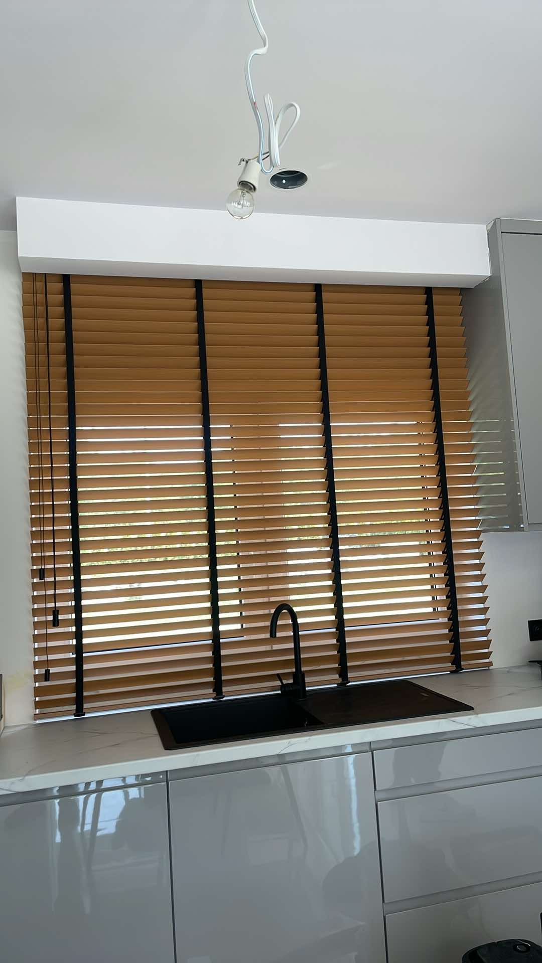 Drewniane żaluzje w kolorze miodowym z czarnymi taśmami w nowoczesnej kuchni z szarymi szafkami i czarnym zlewem. Widoczne niedokończone oświetlenie sufitowe.
