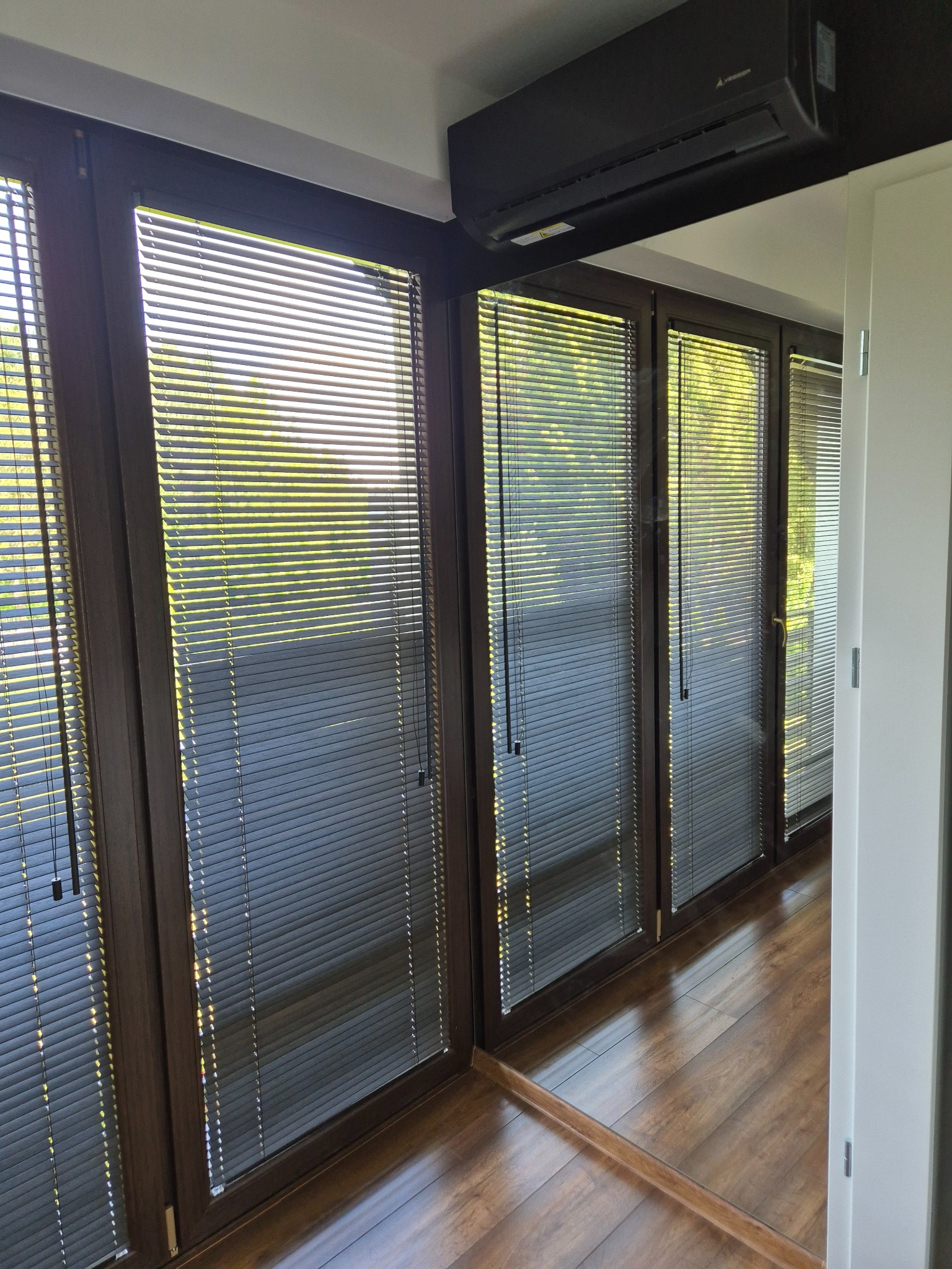 Brązowe okna z aluminiowymi żaluzjami w nowoczesnym wnętrzu. Widoczne refleksy światła na drewnianej podłodze i klimatyzator na suficie.