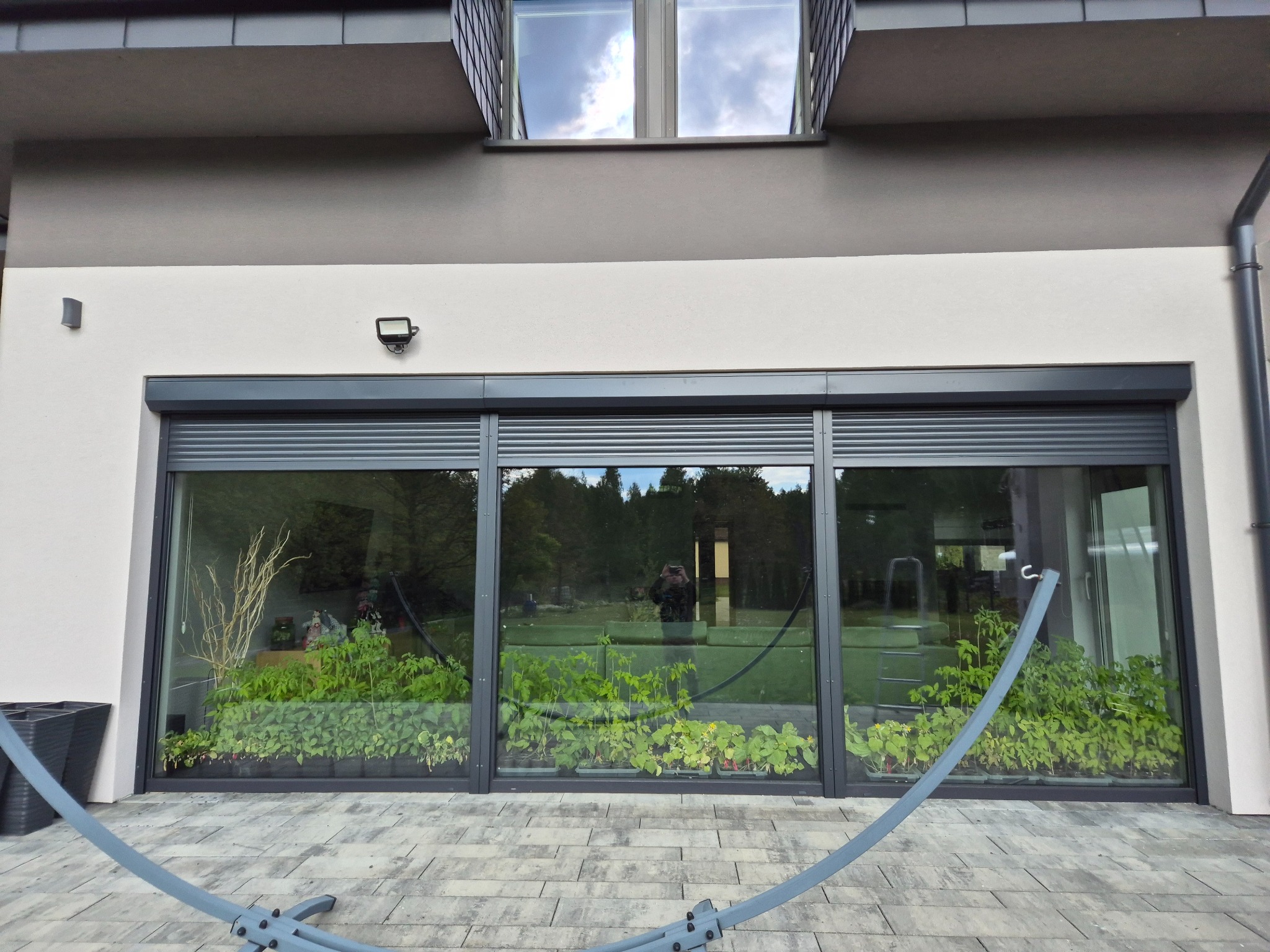 Nowoczesny dom z panoramicznym oknem i częściowo opuszczoną roletą zewnętrzną, widok na rośliny w donicach wewnątrz. Minimalistyczna architektura.