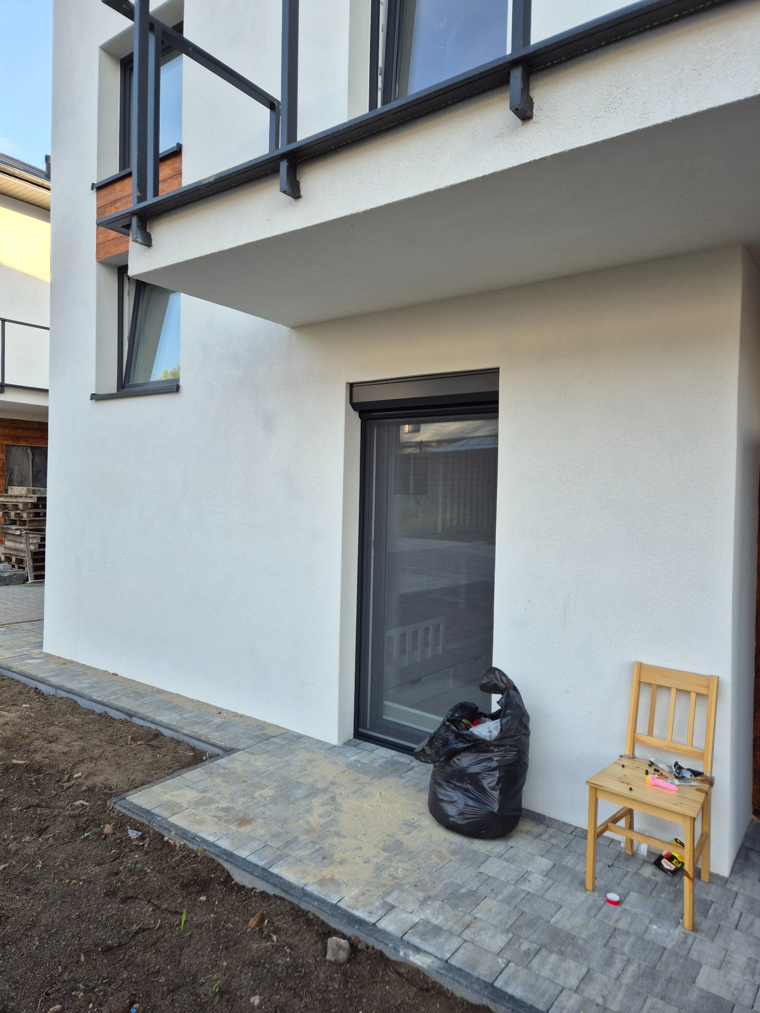 Nowoczesny dom z zamontowaną moskitierą rolowaną w oknie. Obok czarny worek na śmieci i krzesło z narzędziami.