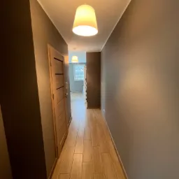 Korytarz z panelami podłogowymi imitującymi drewno, pomalowanymi ścianami w odcieniach szarości i beżu, oświetlony dwoma wiszącymi lampami z abażurami w kształcie ściętego stożka.