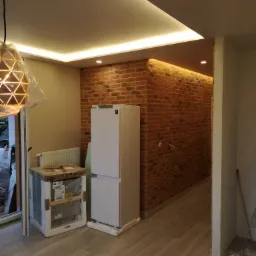 Wnętrze w trakcie prac wykończeniowych: ceglana ściana z podświetleniem LED, nowa lod&oacute;wka w folii obok grzejnika, podłoga imitująca drewno, widoczna puszka z zaprawą i lampa wisząca w osłonie.