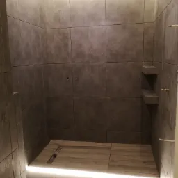 Nowoczesna kabina prysznicowa z szarymi płytkami i drewnopodobną podłogą z odpływem liniowym, oświetlenie LED w suficie i pod podłogą.