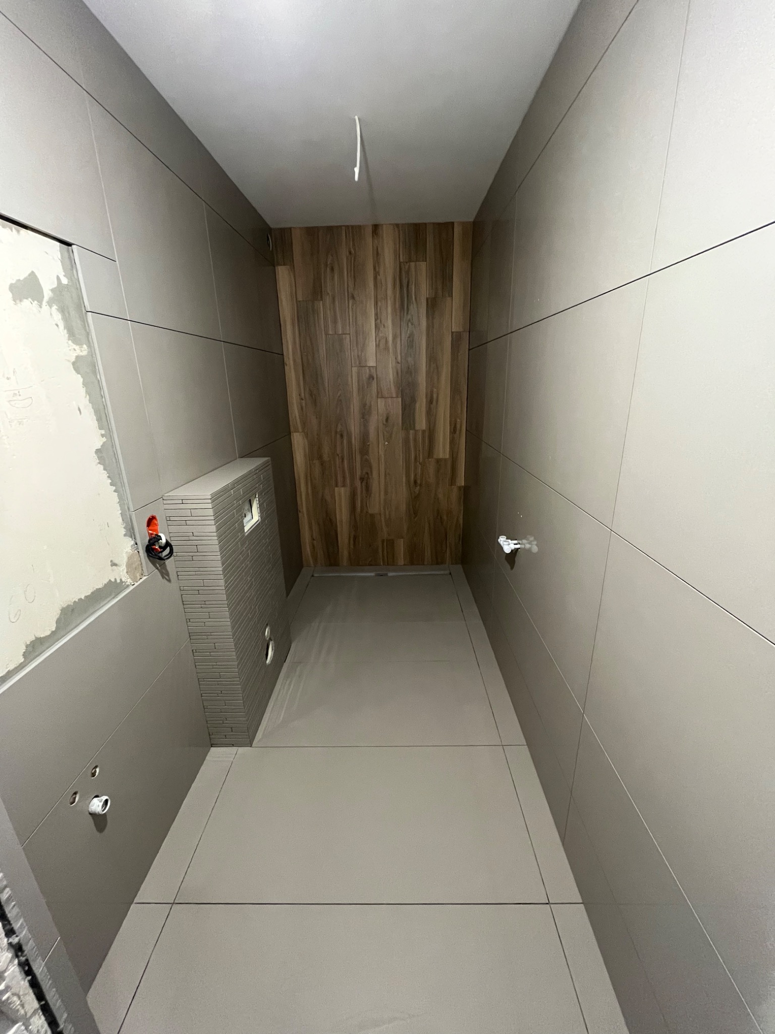 Wnętrze łazienki w trakcie remontu: szare płytki na ścianach i podłodze, drewnopodobna okładzina na jednej ze ścian, zabudowa stelaża WC z płytek, widoczne instalacje hydrauliczne.