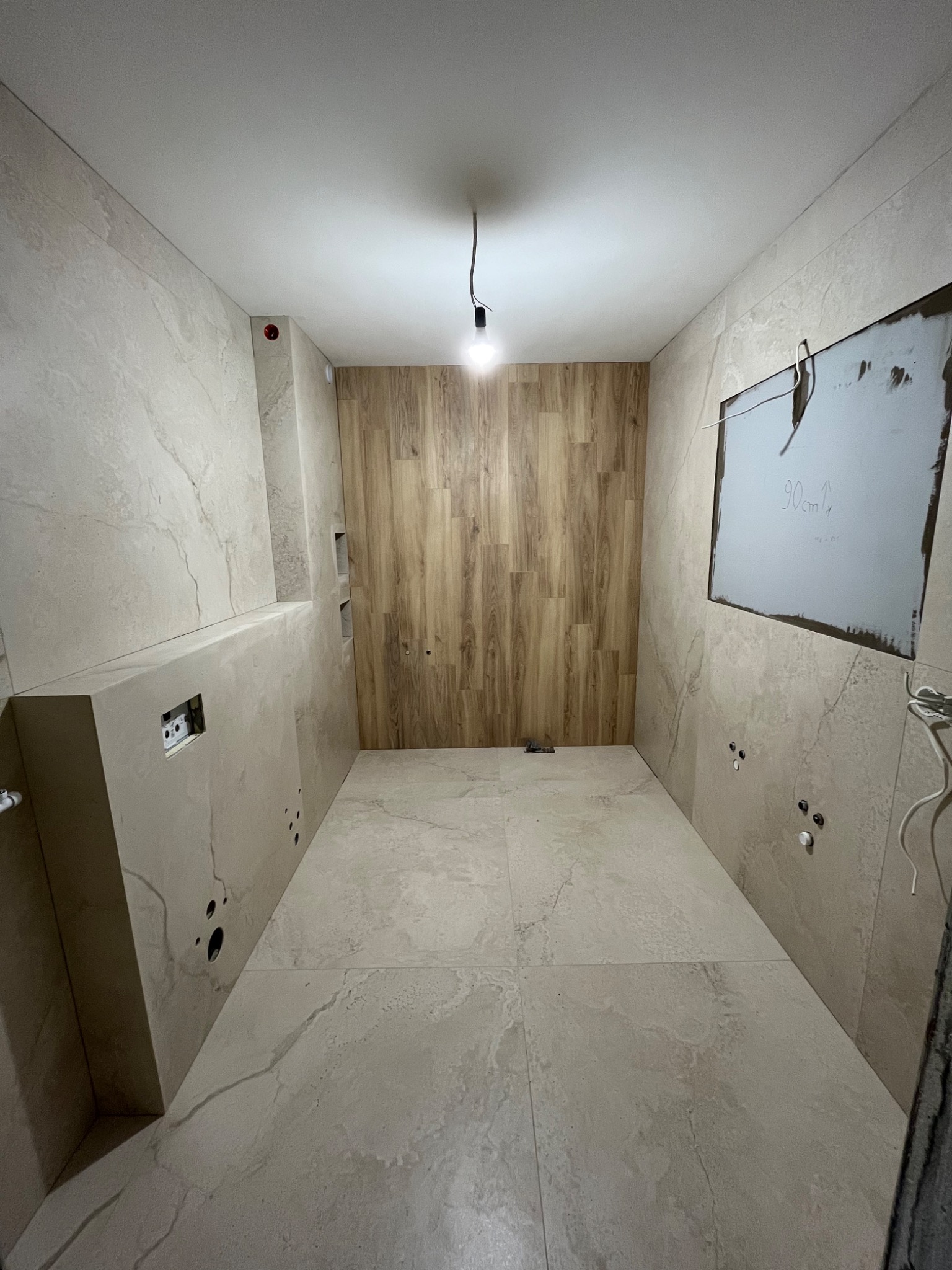 Wnętrze łazienki w trakcie remontu: ściany i podłoga wyłożone płytkami imitującymi marmur, jedna ściana wykończona panelami drewnopodobnymi, widoczne instalacje elektryczne i hydrauliczne...