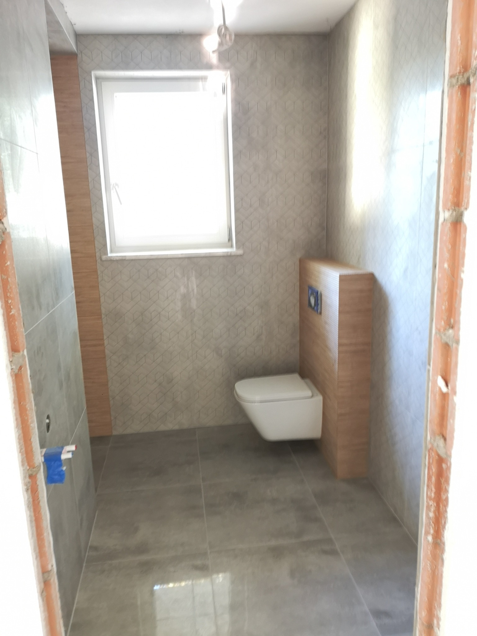 Wnętrze łazienki w trakcie remontu, widoczne szare płytki podłogowe i ścienne z geometrycznym wzorem, wisząca toaleta z zabudową z drewnopodobnej płyty, surowa ściana z cegły w futrynie drzwi.
