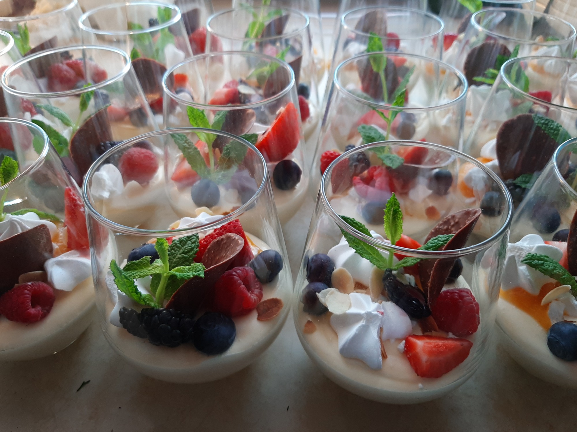 Rząd szklanek wypełnionych deserem z białej czekolady, świeżymi owocami (maliny, borówki, truskawki, jeżyny), bezami, listkami mięty i płatkami czekolady.