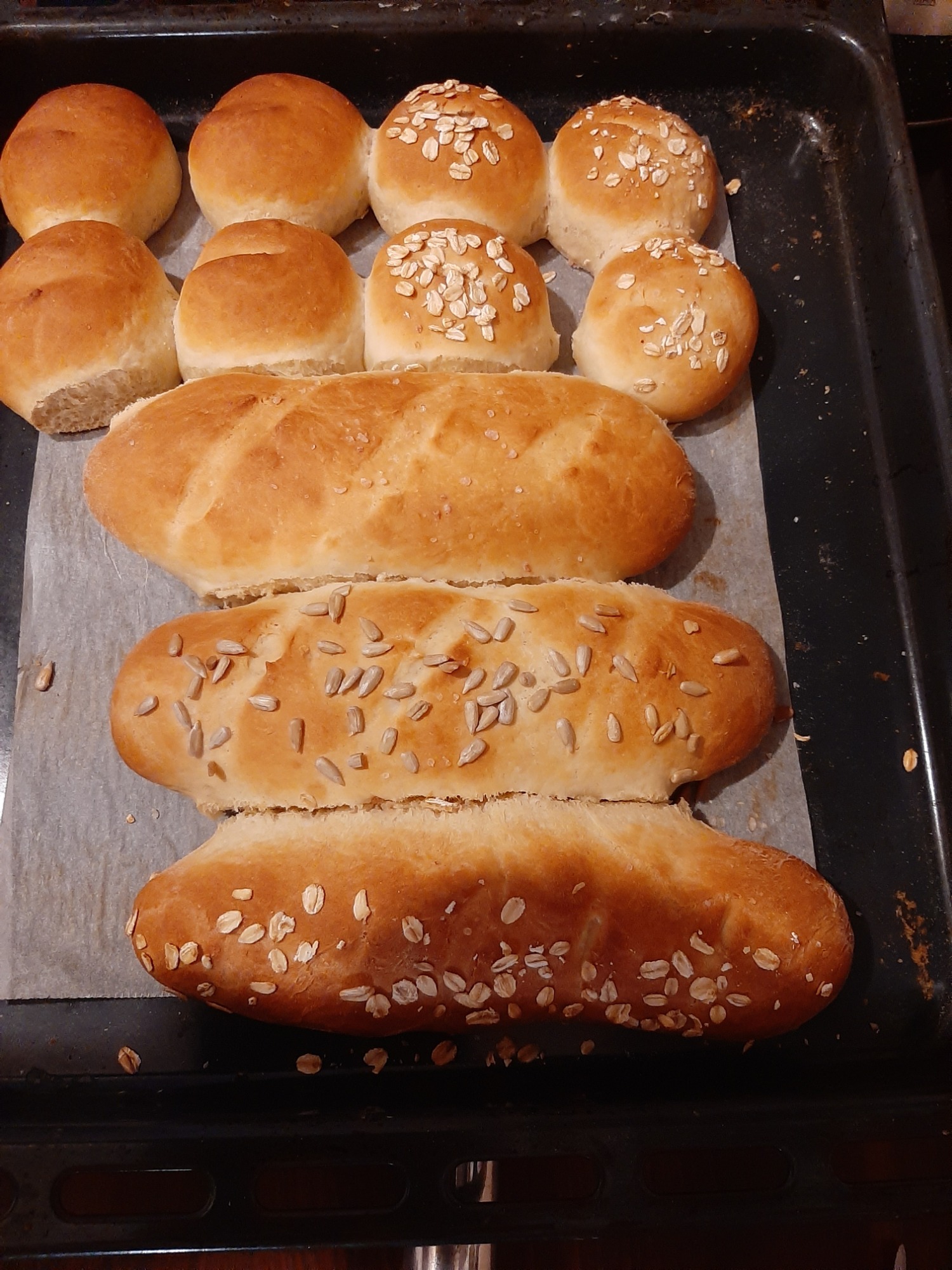Świeżo upieczone bułki i bagietki na blasze wyłożonej papierem do pieczenia, część posypana płatkami owsianymi, a część ziarnami słonecznika.