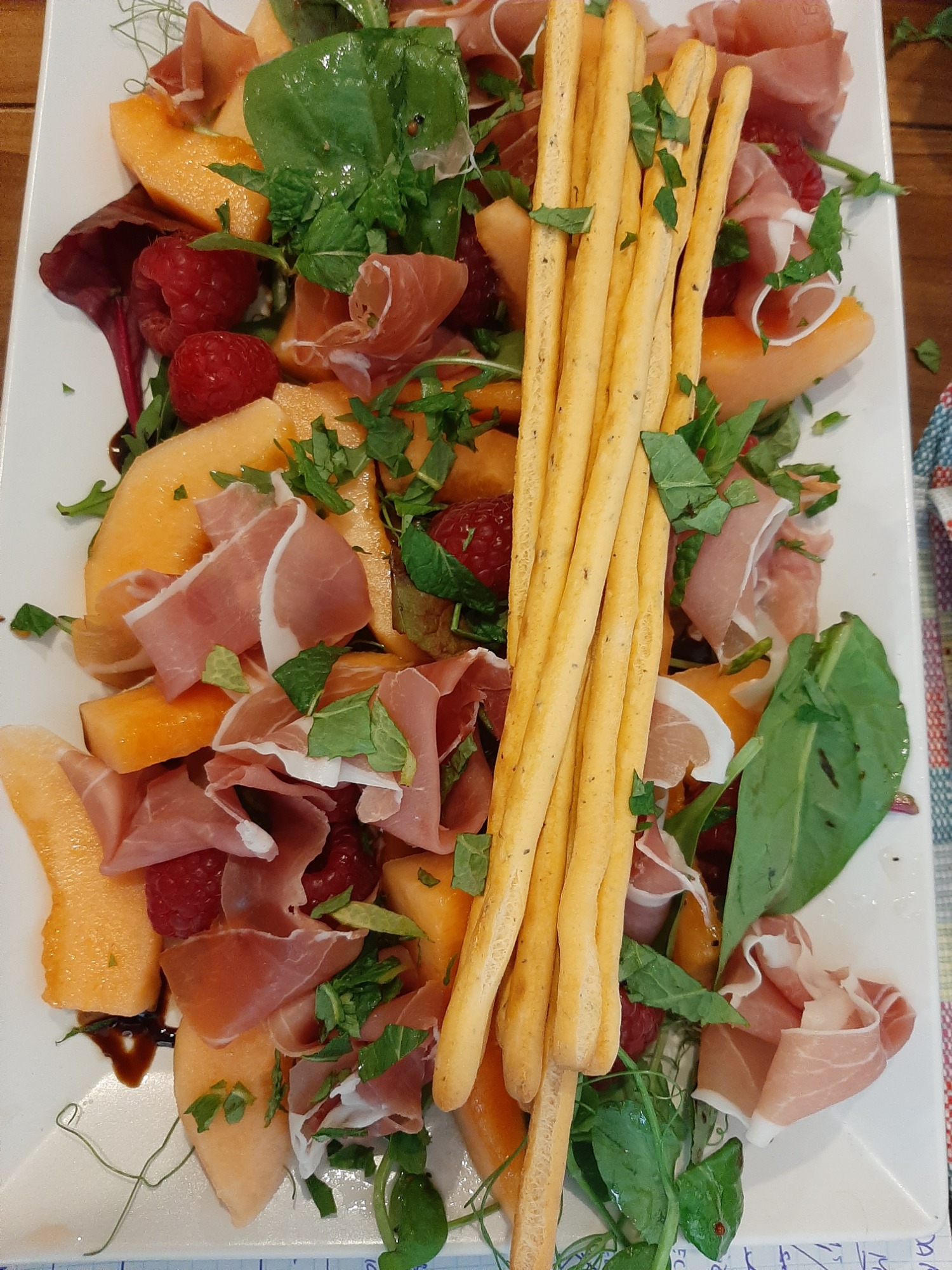 Apetyczna sałatka na białym talerzu: kawałki melona, szynka parmeńska, maliny, grissini i listki mięty, skropione ciemnym sosem.