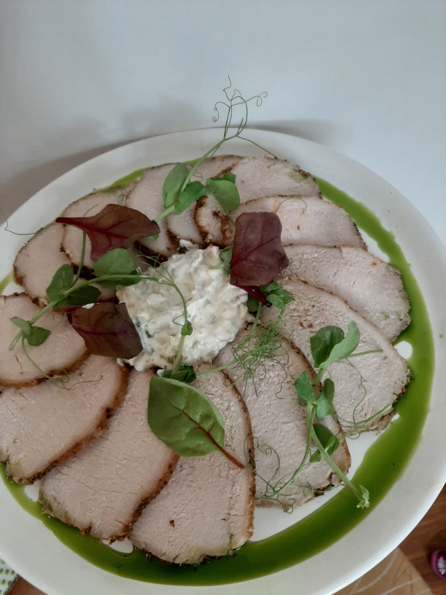 Plasterki pieczonego schabu z sosem tatarskim i szczypiorkową oliwą, dekorowane zielonymi pędami i listkami na białym talerzu z zielonym sosem.