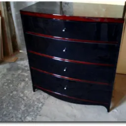 Gięta komoda z czterema szufladami w czarnym, wysokim połysku, z mahoniowymi akcentami, sfotografowana w warsztacie na tle materiałów budowlanych.